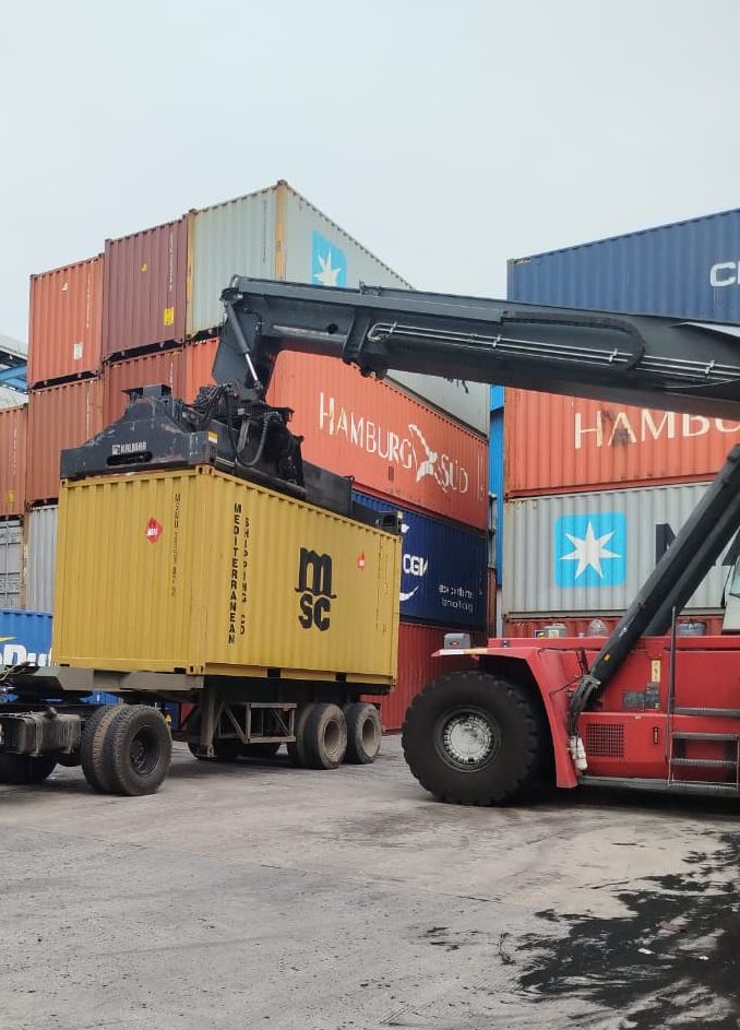 Alat berat reach stacker memuat kontainer ke atas armada truk logistik USLOGT di area depo untuk pengiriman kargo yang efisien.